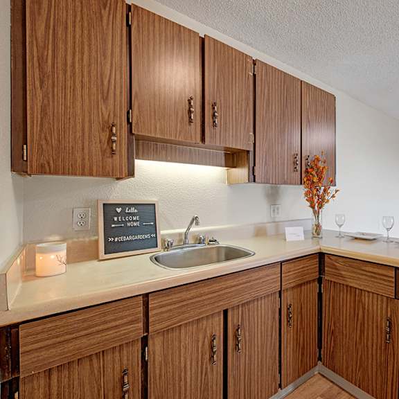 cozy kitchen here at cedar gardens apartment homes with wooden cabinets, a small sink, and a lighted backsplash. a welcoming sign, candle, and two place settings add warmth.
