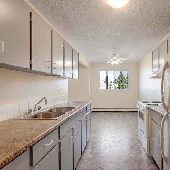 narrow kitchen here at cedar gardens apartment homes with light gray cabinets, white appliances, and granite countertops. a window at the end overlooks trees, creating an airy atmosphere.