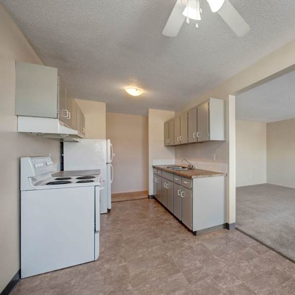 simple kitchen here at cedar gardens apartment homes with white appliances, including a stove and refrigerator. gray cabinets line the walls. adjacent is a carpeted living room, creating an open and neutral space.