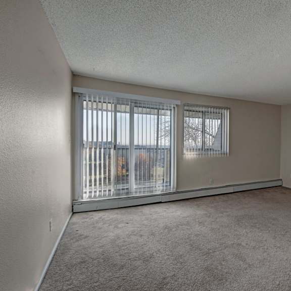 spacious, empty living room here at cedar gardens apartment homes with beige walls and carpet. large sliding glass doors and window with vertical blinds reveal a view of trees and grass outside. quiet and serene.