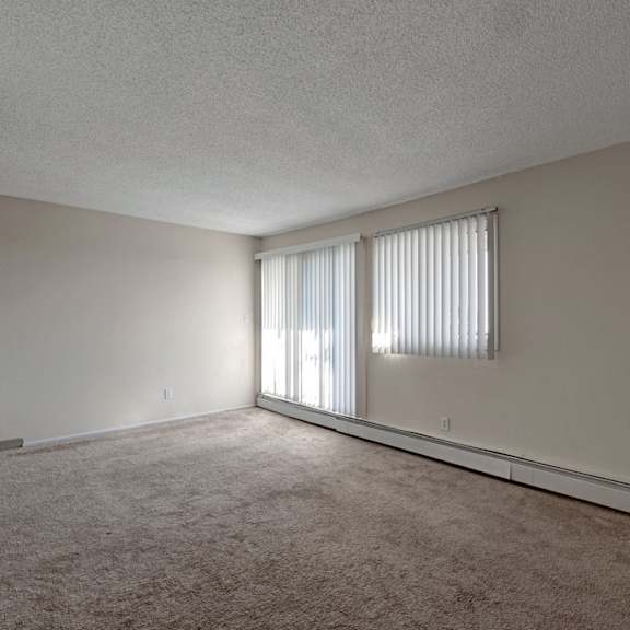 empty room here at cedar gardens apartment homes with beige carpet and white walls, featuring two vertical blind-covered windows letting in soft light, creating a calm, minimalist atmosphere.
