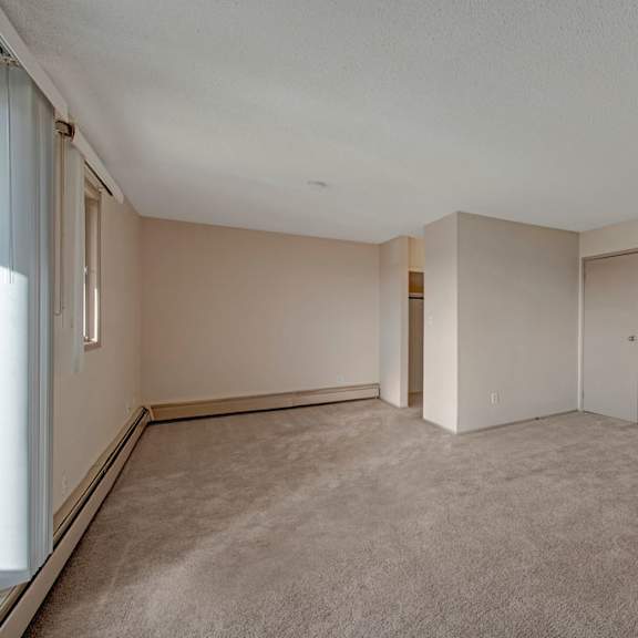 an empty room here at cedar gardens apartment homes with beige walls and carpet, featuring a closed door, small window on the left, and open closet. soft lighting creates a calm atmosphere.