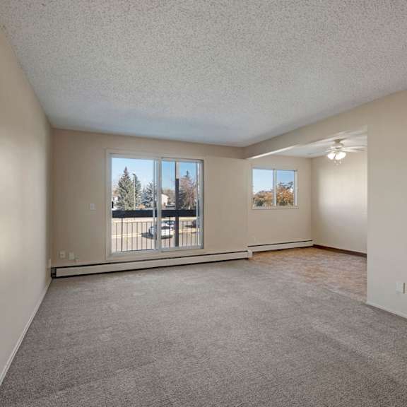 spacious, empty living room here at cedar gardens apartment homes with beige walls and carpet, large window showing trees outside. adjacent dining area with a ceiling fan and tiled floor.