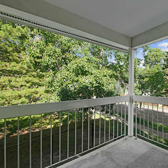 covered balcony here at gateway gardens apartment homes with metal railing overlooks lush green trees and a quiet street. the scene feels serene and inviting under a clear blue sky.