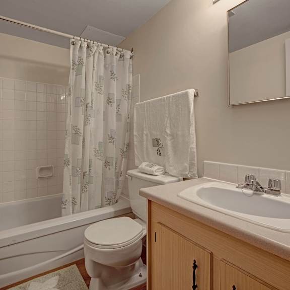 small bathroom here at gateway gardens apartment homes with a bathtub and shower curtain on the left, toilet in the center, and wooden cabinet with sink and mirror on the right. bright and clean atmosphere.