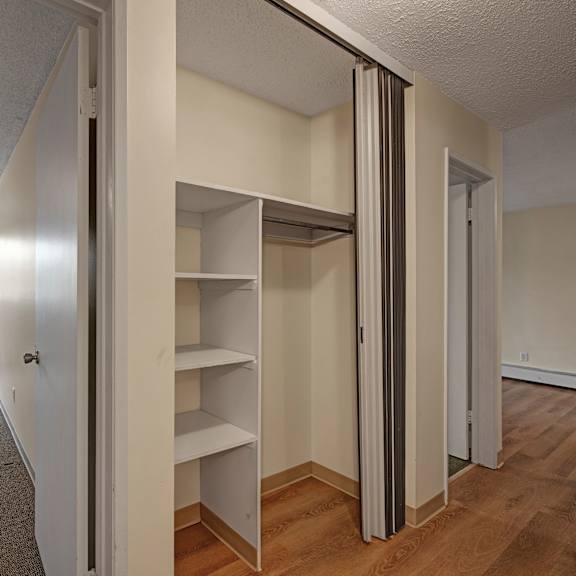 empty room here at gateway gardens apartment homes with beige walls and carpet, illuminated by a square ceiling light. open closet with shelves on the right. window with vertical blinds reveals greenery outside.