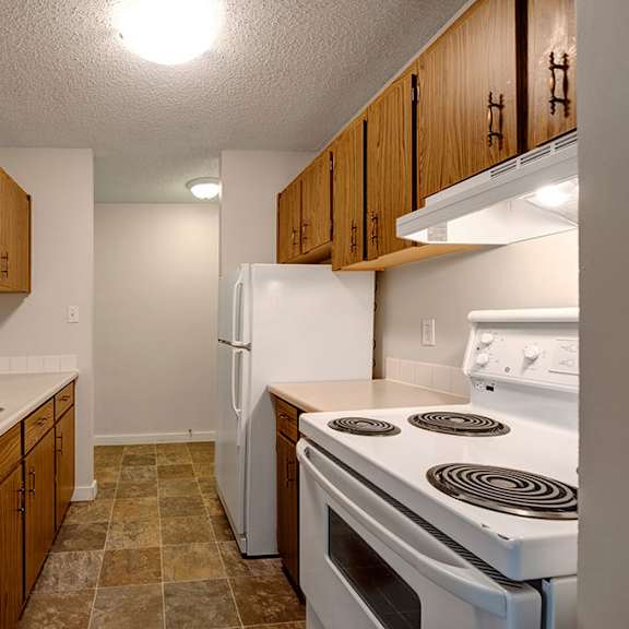 narrow kitchen here at gateway gardens apartment homes with wooden cabinets, a white stove, and fridge. a stainless steel sink on the left; tile floor and bright overhead lighting.