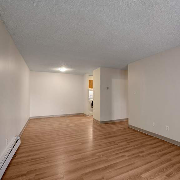 empty living room here at gateway gardens apartment homes with light brown wood flooring and beige walls, illuminated by a ceiling light. an open doorway leads to a small kitchenette. minimalistic and clean.