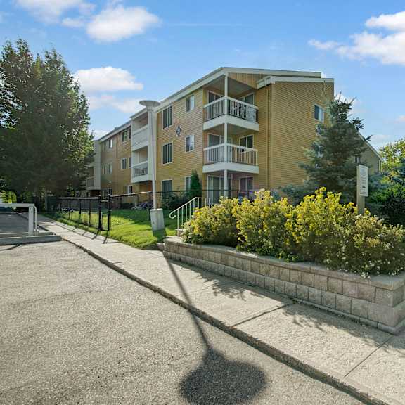 three-story beige gateway gardens apartment homes building under a blue sky, surrounded by trees and shrubs. a sidewalk and driveway are in the foreground. peaceful atmosphere.