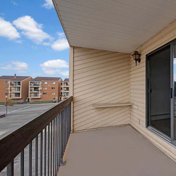 apartment balcony here at gateway square apartment homes with beige siding and sliding glass doors, overlooking nearby residential buildings and greenery. clear blue sky with fluffy clouds.