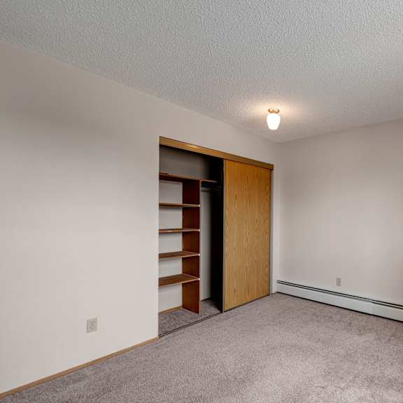 empty bedroom here at gateway square apartment homes with beige walls, a small ceiling light, and carpeted floor. open closet with shelves on the left, creating a clean, neutral atmosphere.