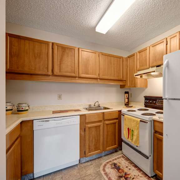 cozy kitchen here at gateway square apartment homes with wooden cabinets, white appliances, and beige countertops. features include a fridge, stove, dishwasher, and decorative rug. simple and neat.