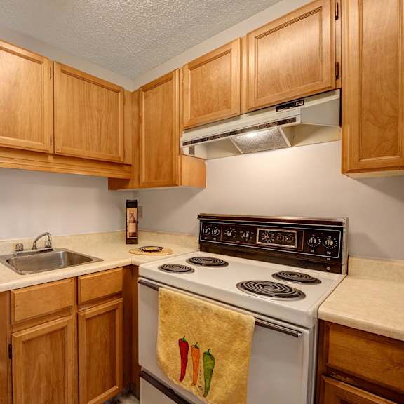 cozy kitchen here at gateway square apartment homes with light wood cabinets, an electric stove, and beige countertops. a hand towel with chili peppers hangs on the oven, creating a warm, inviting feel.