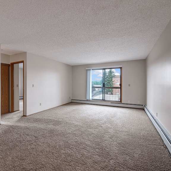 spacious empty living room here at gateway square apartment homes with beige carpet, white walls, and large window allowing natural light. open doorway leads to another room. tranquil ambiance.