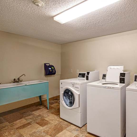 laundry room here at lakewood heights apartment homes with two white top-load washing machines and a front-load dryer. a utility sink with a faucet is on the left. beige walls and tiled floor.