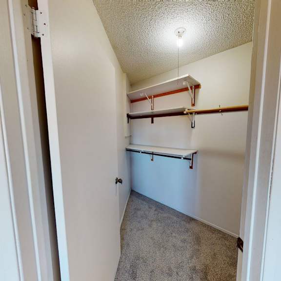 a small walk-in closet here at lakewood heights apartment homes with beige carpet, white walls, and a single ceiling light. two wooden rods and white shelves for storage are visible.