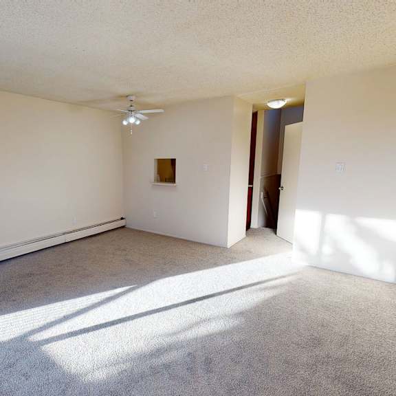 empty bedroom here at lakewood heights apartment homes with beige walls and gray carpet, illuminated by sunlight. features include a ceiling fan, small wall cutout, and an open door. minimalist and bright atmosphere.