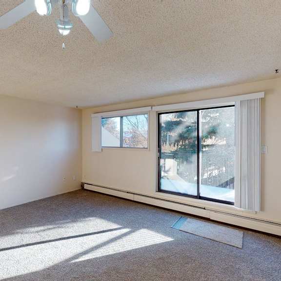 empty room here at lakewood heights apartment homes with beige walls, carpeted floor, and ceiling fan. large sliding glass doors and a window let in sunlight, casting shadows across the room.
