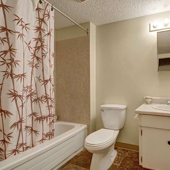 compact bathroom here at lakewood heights apartment homes with beige walls, featuring a bamboo-patterned shower curtain, white toilet, and sink with a mirror above. soft, warm lighting.