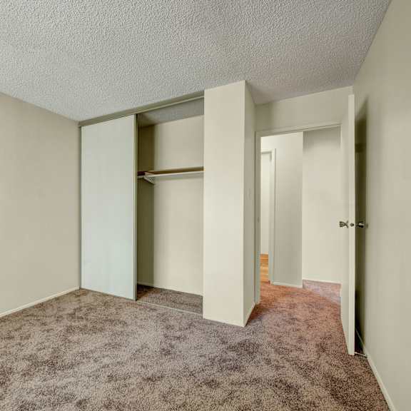 empty bedroom here at lakewood heights apartment homes with beige walls and carpet, featuring an open closet with sliding doors. a partially open door reveals a hallway, creating a calm, neutral tone.