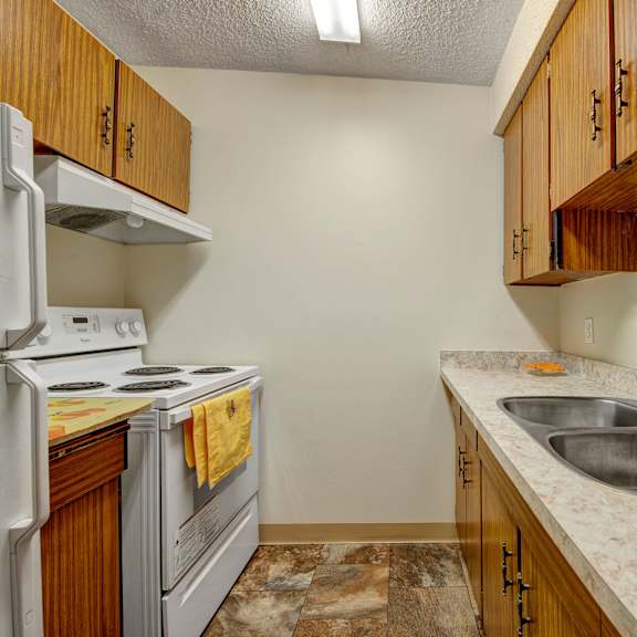 compact kitchen here at lakewood heights apartment homes with white appliances and brown wooden cabinets. marble countertops with a double sink and a yellow towel on the stove, creating a cozy feel.