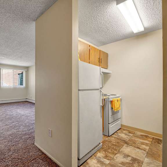 spacious empty room here at lakewood heights apartment homes with beige carpet and two large windows with vertical blinds. adjacent kitchen with white appliances and yellow towel. bright and minimal.