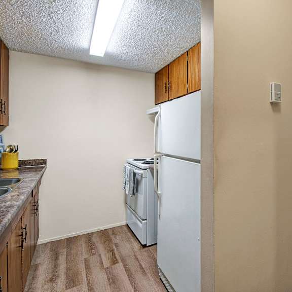 compact kitchen here at lakewood heights apartment homes with wood cabinets and a white stove, adjacent to a bright living room with beige carpet and large window showing a grassy view. cozy atmosphere.