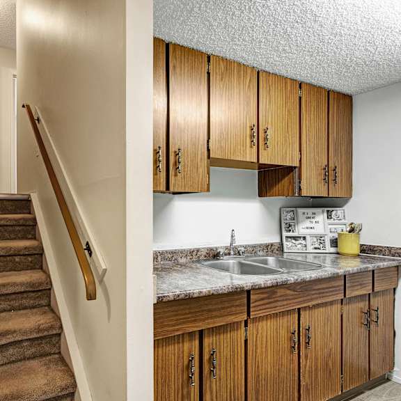 carpeted stairs lead to an upper floor beside a small kitchen with wood cabinets and a marble countertop here at lakewood heights apartment homes. the tones are warm and cozy.