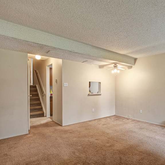 empty living room here at lakewood heights apartment homes with beige walls and carpet, ceiling fan, and a small window opening to a kitchen area. stairs are visible in the background.