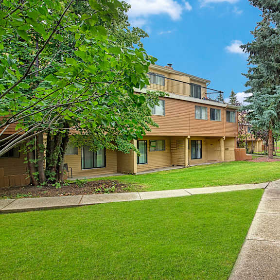 spacious green backyard here at lakewood heights apartment homes with a beige two-story house, surrounded by lush trees and manicured lawns. a winding concrete path leads through the serene landscape.