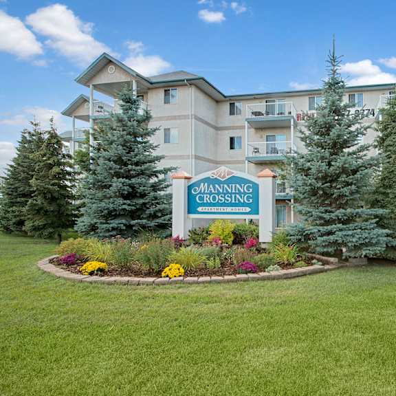 apartment complex with beige exterior and balconies on four floors. sign reads "manning crossing apartment homes" surrounded by manicured garden and trees.