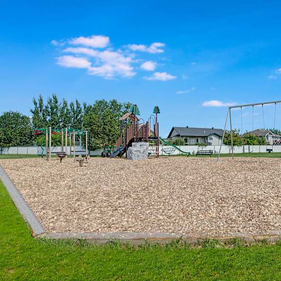 playground here at sol terrace apartment homes with swings and climbing set, surrounded by wood chips and grass. houses and trees in the background under a bright blue sky.