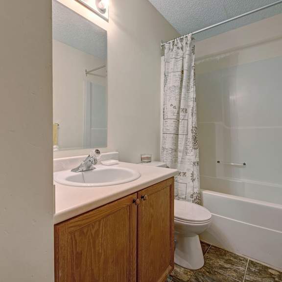 small bathroom here at sol terrace apartment homes with wood cabinet, sink, and toilet. a white shower curtain with decorative patterns hangs over a bathtub. light tones create a clean, simple look.
