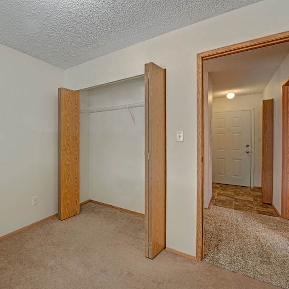 empty room here at sol terrace apartment homes with beige carpet, open wooden closet doors revealing a shelf, and an entrance leading to a lit hallway with a closed white door. simple and neutral atmosphere.