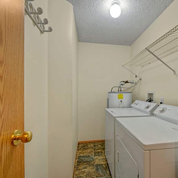 laundry room here at sol terrace apartment homes with a washing machine, dryer, and water heater. an overhead shelf is mounted on the right wall. the room has beige walls and tiled floor.