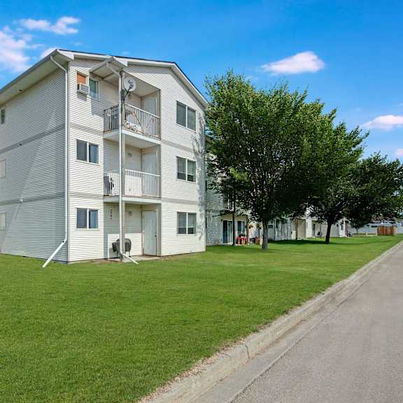 three-story sol terrace apartment building with white siding, surrounded by green lawns and vibrant trees under a bright blue sky. a parked blue suv is visible.