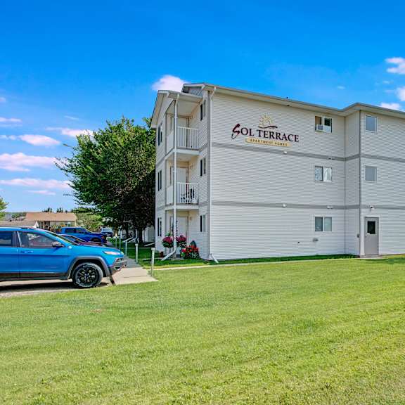 four-story beige apartment building, labeled "sol terrace," with well-maintained lawn, trees, and a parked blue suv under a bright blue sky.