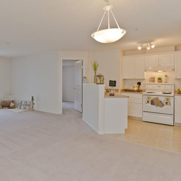 spacious living area here at somerset pointe apartment homes with beige carpet, sliding glass doors on the left, cozy decor including plants and pillows, and a bright, white kitchen on the right.