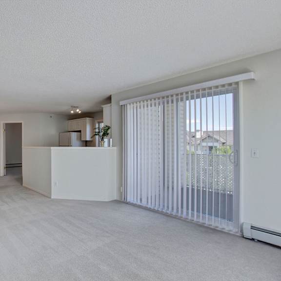 spacious, empty living room here at somerset pointe apartment homes with light gray carpet, white walls, and vertical blinds on a sliding glass door leading to a balcony. modern and airy feel.
