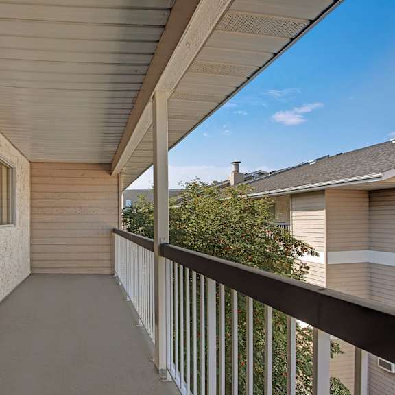 covered balcony here at sterling place apartment homes with beige stucco walls, sliding glass door, and black railing. overlooks a tree and neighboring beige building under a clear blue sky.