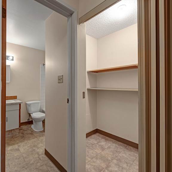 a narrow hallway here at sterling place apartment homes leading to a bathroom with a toilet, white tiles, and a small vanity with a light above, and a closet with empty shelves on the right.