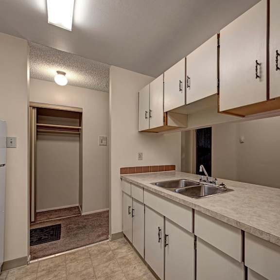 narrow kitchen here at sterling place apartment homes with beige cabinets and countertops, a double stainless steel sink, and a fridge. adjacent room visible through a doorway, creating a cozy feel.