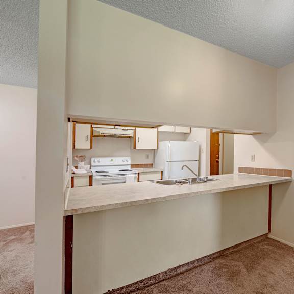 small, unfurnished kitchen here at sterling place apartment homes with beige walls and carpet. features a countertop with sink, white appliances, and an open layout, creating a simple, cozy vibe.