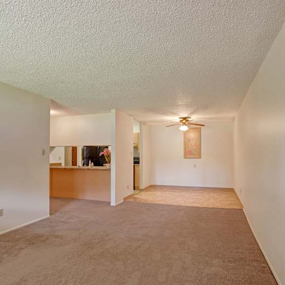 empty living room here at sterling place apartment homes with beige carpeting and white walls, leading to an open kitchen area with a ceiling fan. soft lighting and minimal decor give a neutral, calm feel.