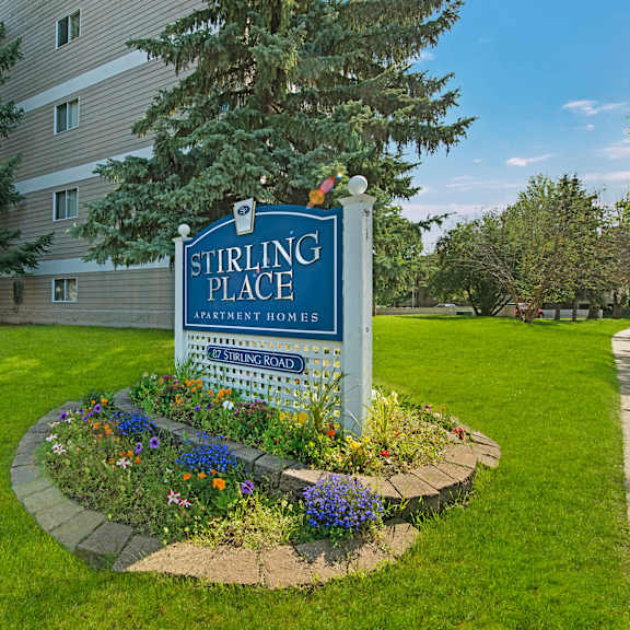 sign for stirling place apartment homes, surrounded by colorful flowers and lush grass, with a tall building and evergreen trees in the background.