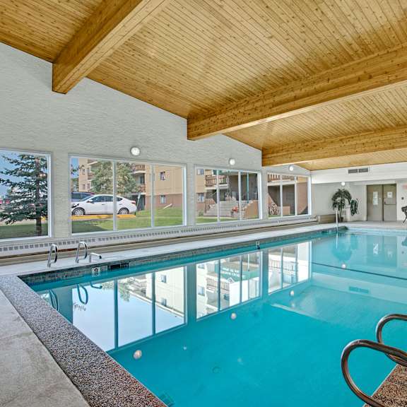 indoor swimming pool here at wyndham crossing apartment homes with clear blue water, surrounded by large windows showing outdoor greenery. wooden ceiling and deck chairs create a serene atmosphere.