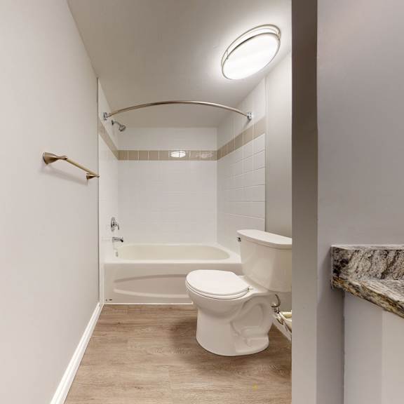 narrow bathroom here at wyndham crossing apartment homes with white bathtub, shower, and toilet. beige tiles line the tub area. granite countertop visible. minimalist and clean design.