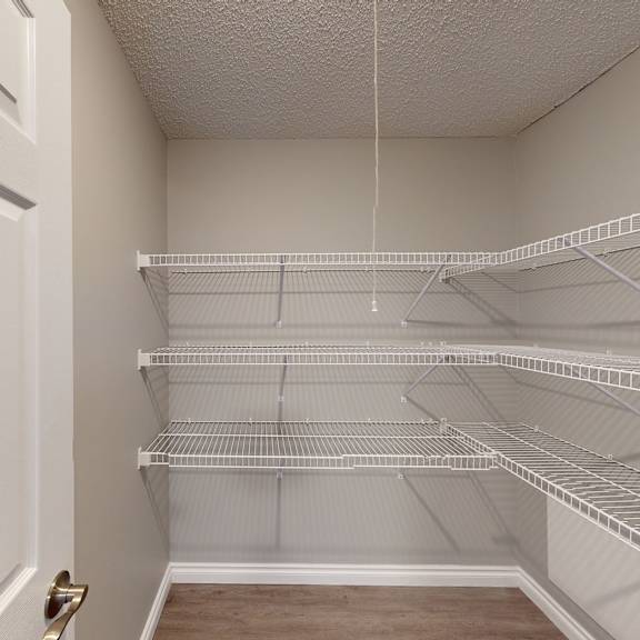 a small, empty closet here at wyndham crossing apartment homes with light gray walls, featuring three rows of white wire shelving on the right. the door is partially open, revealing a wooden floor.