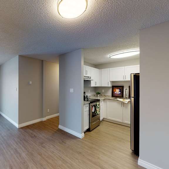 modern apartment interior here at wyndham crossing apartment homes with a spacious living area featuring large windows, wood flooring, and neutral walls. the kitchen has white cabinets and stainless steel appliances, under bright lighting. the scene feels open and inviting.