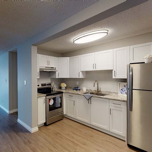 modern kitchen and living area here at wyndham crossing apartment homes with wood floors, stainless steel appliances, white cabinets, and large window with vertical blinds overlooking a snowy scene.